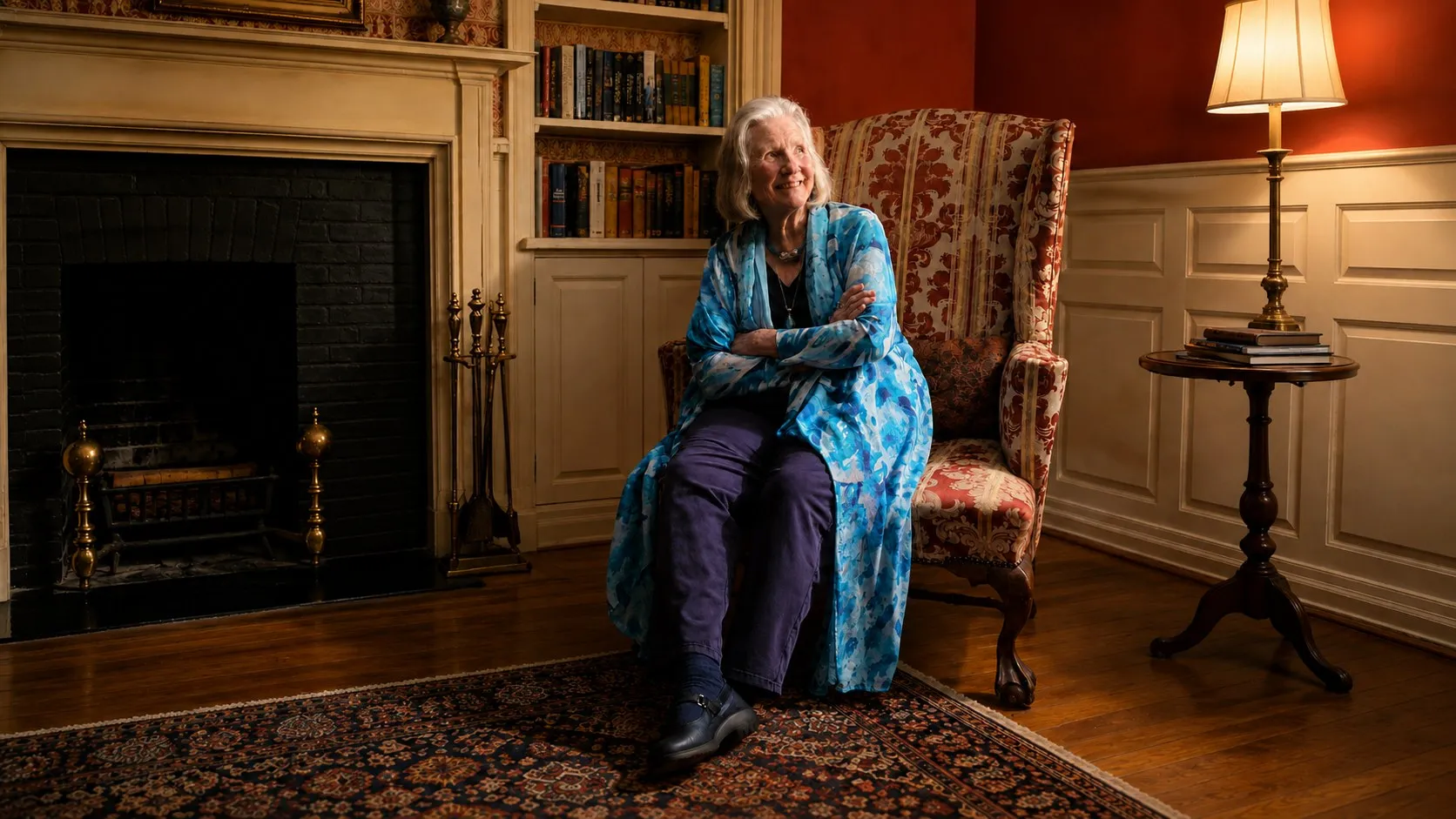 Jan Carlberg seated in a wing chair beside a fireplace and bookshelves